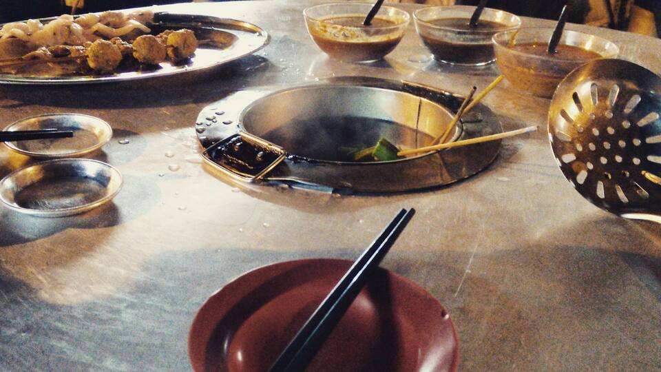 Ein Tisch mit verschiedenen Speisen, darunter eine rote Schüssel mit Stäbchen, eine Metallschüssel mit Deckel und mehrere kleine Schalen mit Soßen.