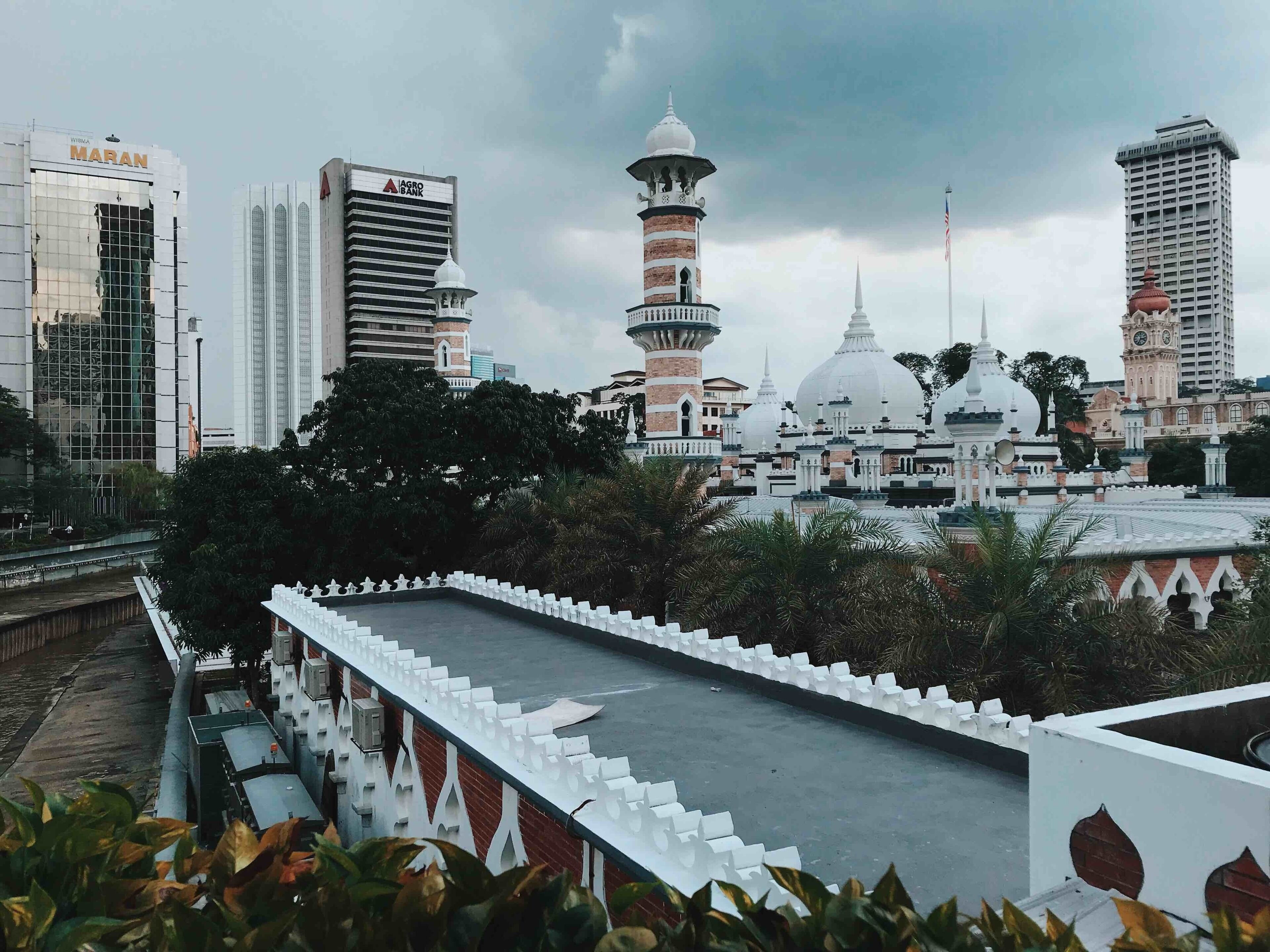 A mosque with a minaret and dome, surrounded by modern buildings and a white fence.