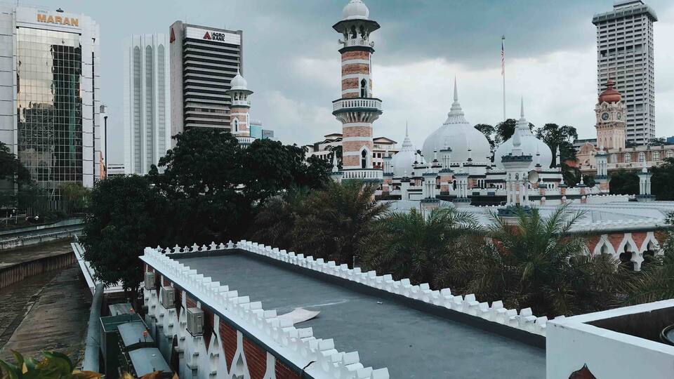 A mosque with a minaret and dome, surrounded by modern buildings and a white fence.