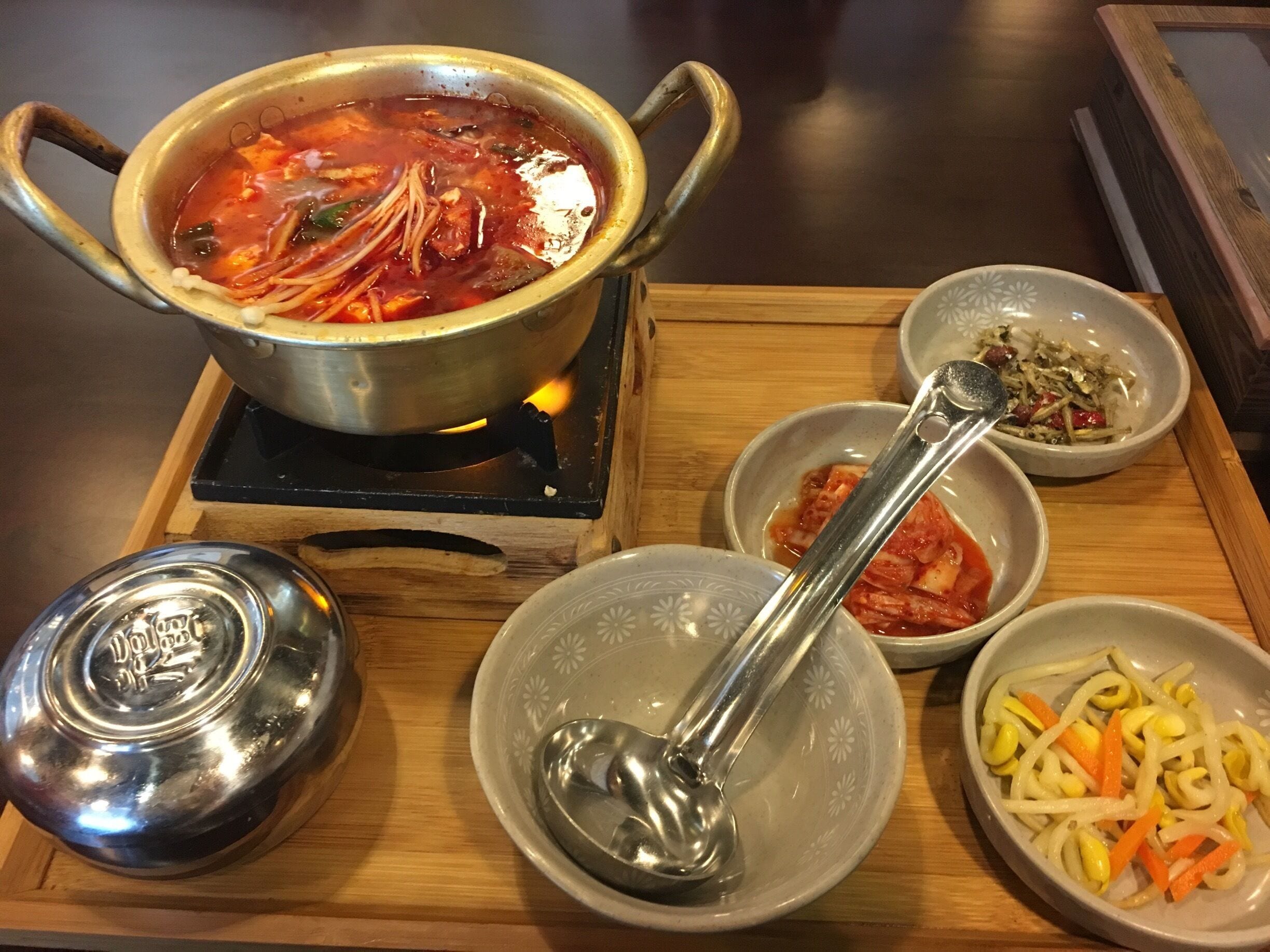 Una olla de sopa sobre la estufa, un tazón de vegetales cortados, un tazón de kimchi y una tapa de metal sobre una bandeja de madera.
