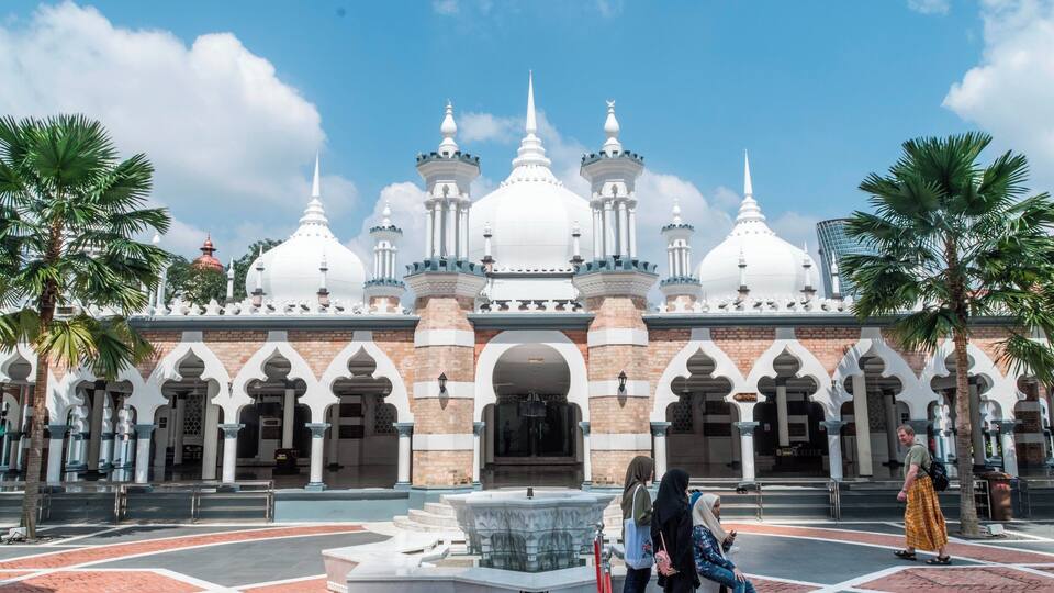 A large white mosque with multiple domes and minarets, surrounded by palm trees and a courtyard with a fountain.