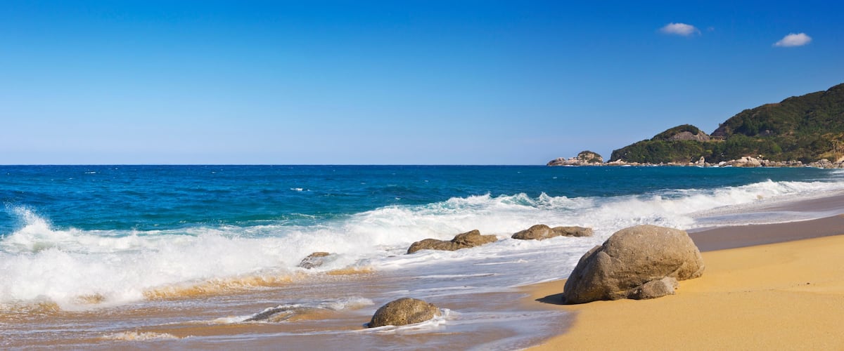 A sandy beach with waves, large rocks, and a mountainous backdrop.