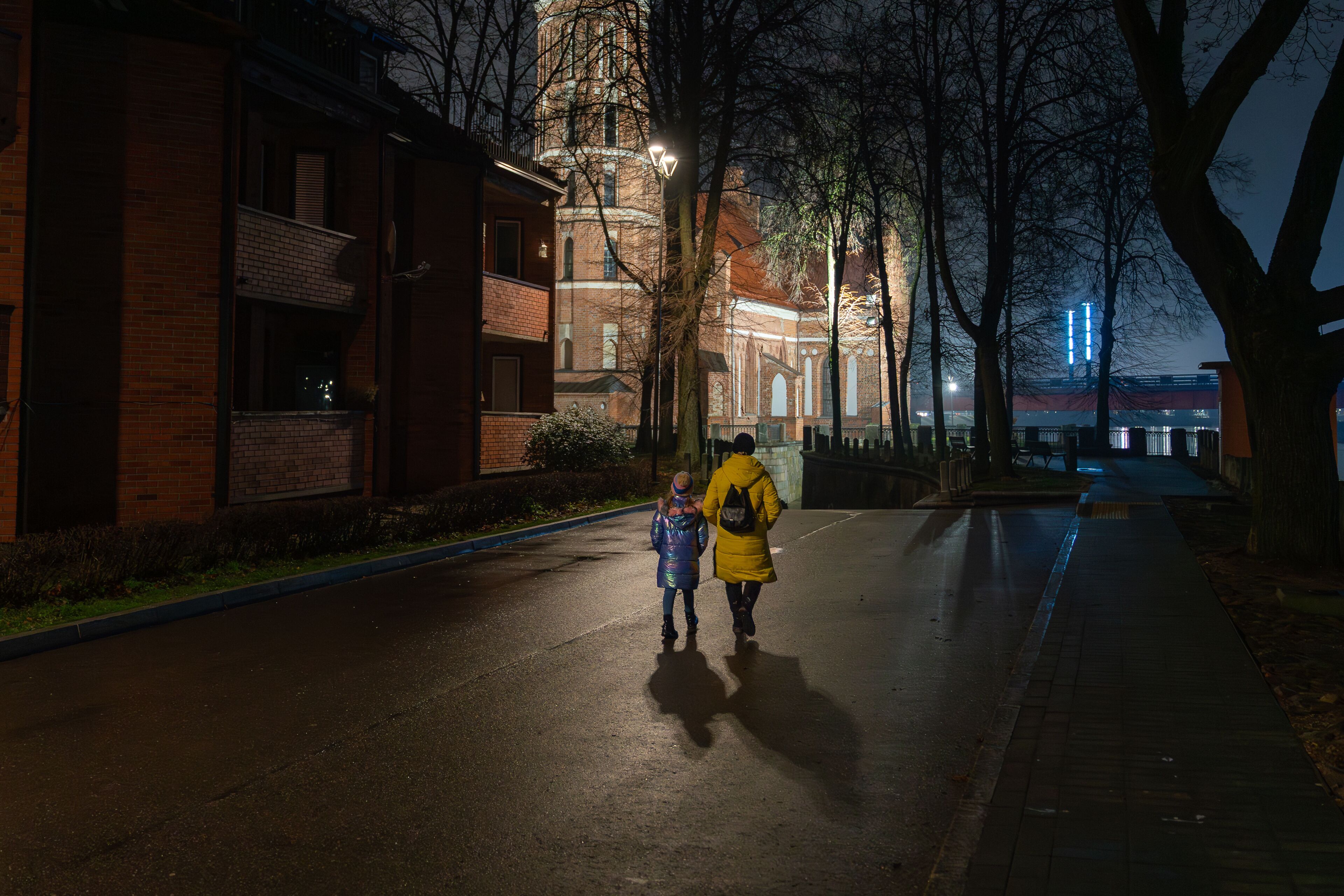 Mother and daughter walking at night toward an illuminated brick church in Kaunas, a festive winter street with wet reflections.