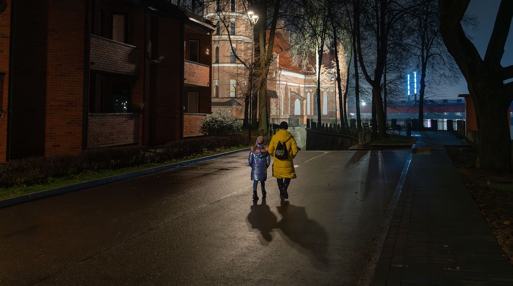 Mother and daughter walking at night toward an illuminated brick church in Kaunas, a festive winter street with wet reflections.