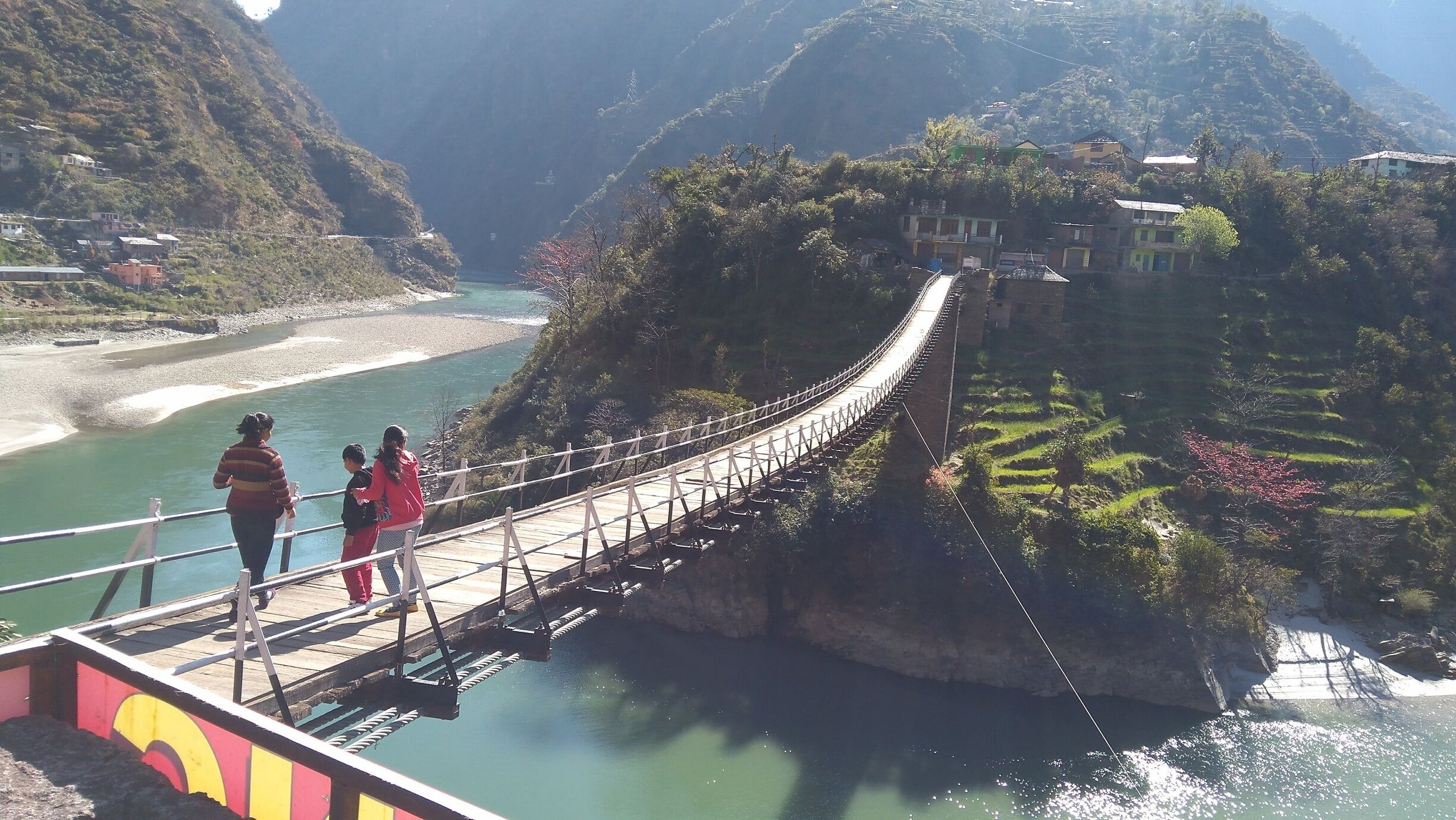 A suspension bridge over a river with terraced fields and houses on the hillside.