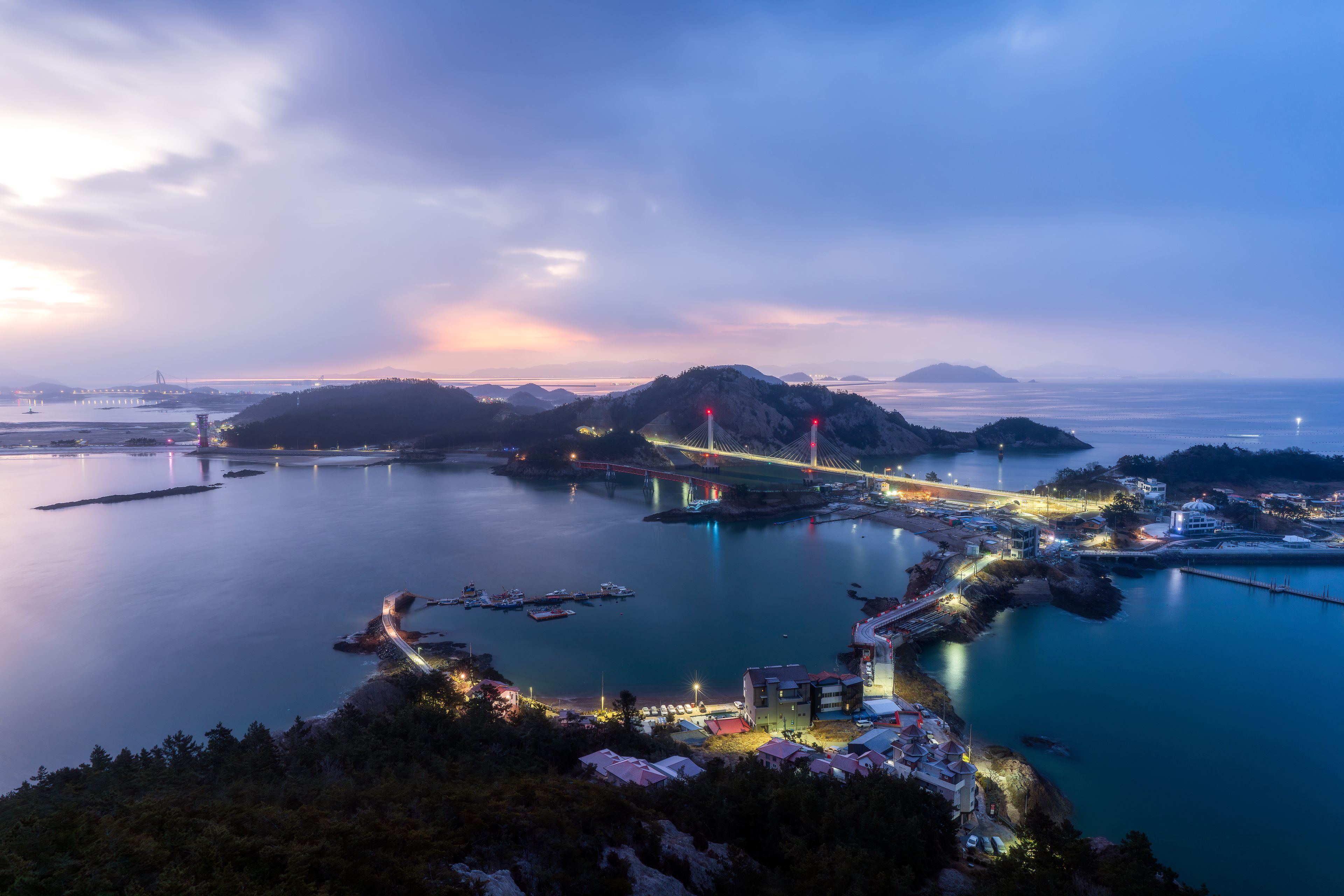 A coastal cityscape at dusk with illuminated bridges, a calm sea, and distant mountains.