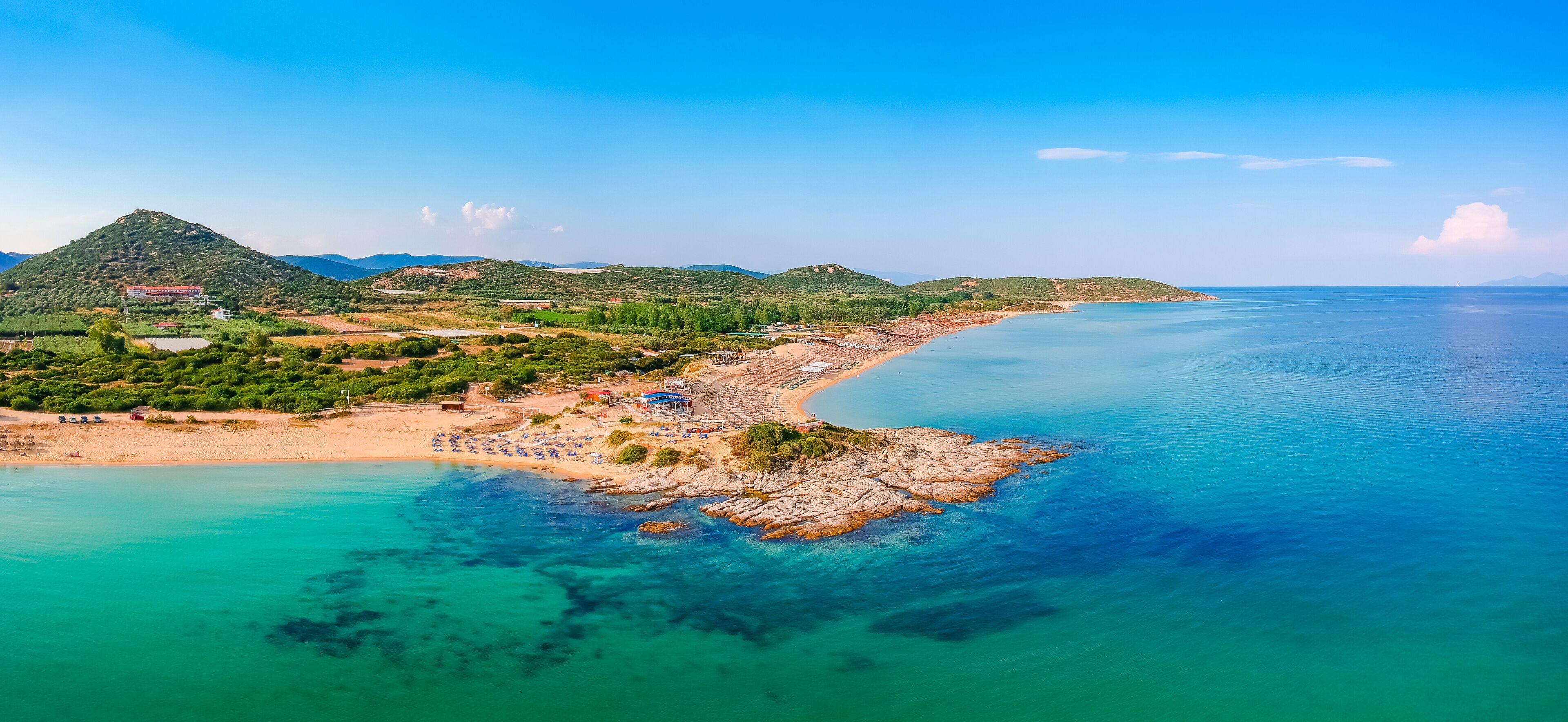 A coastal landscape with a sandy beach, clear turquoise waters, and green hills in the background.