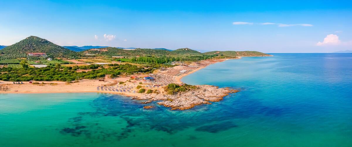 A coastal landscape with a sandy beach, clear turquoise waters, and green hills in the background.