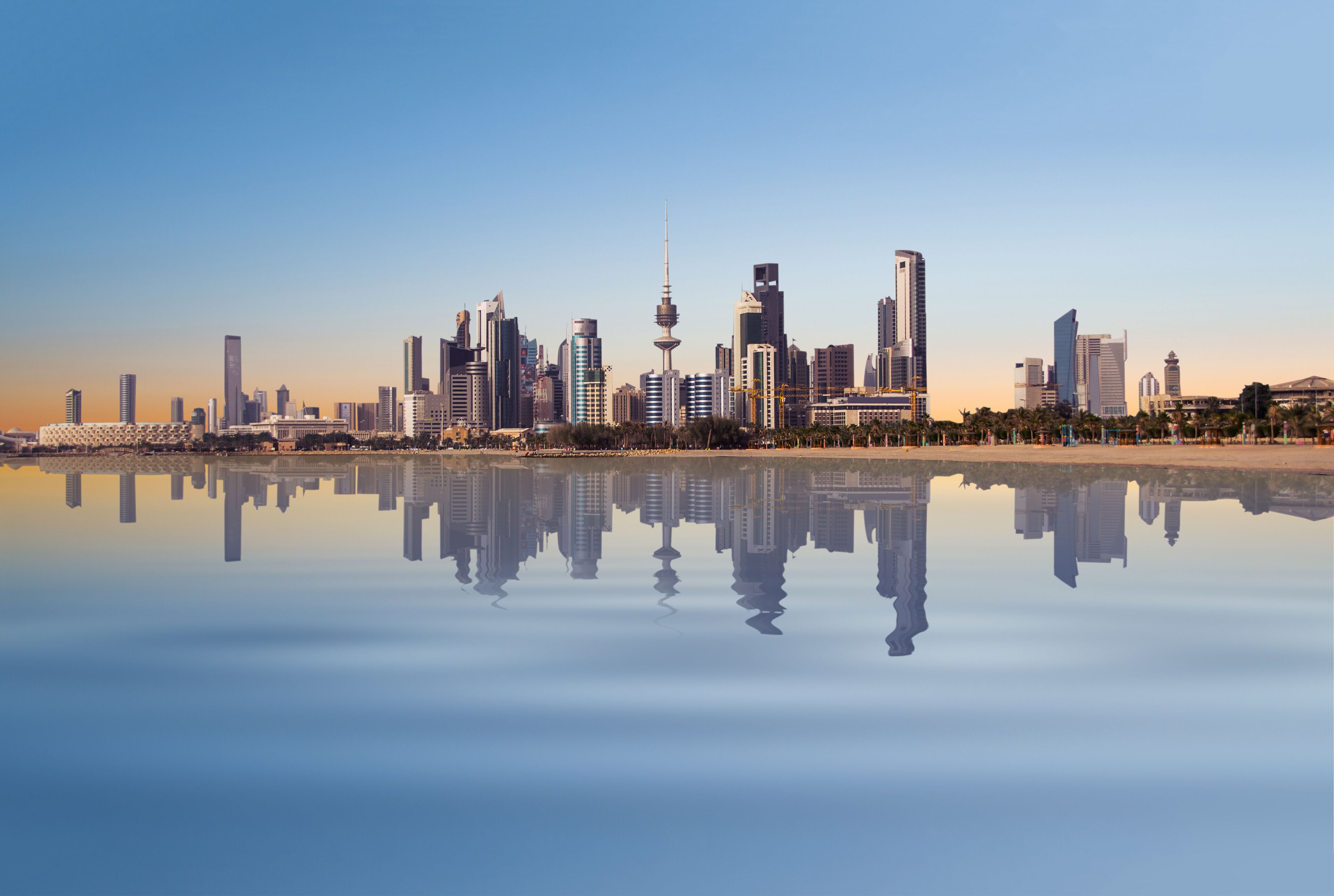 A city skyline reflected in calm water, including a prominent tower and several high-rise buildings.