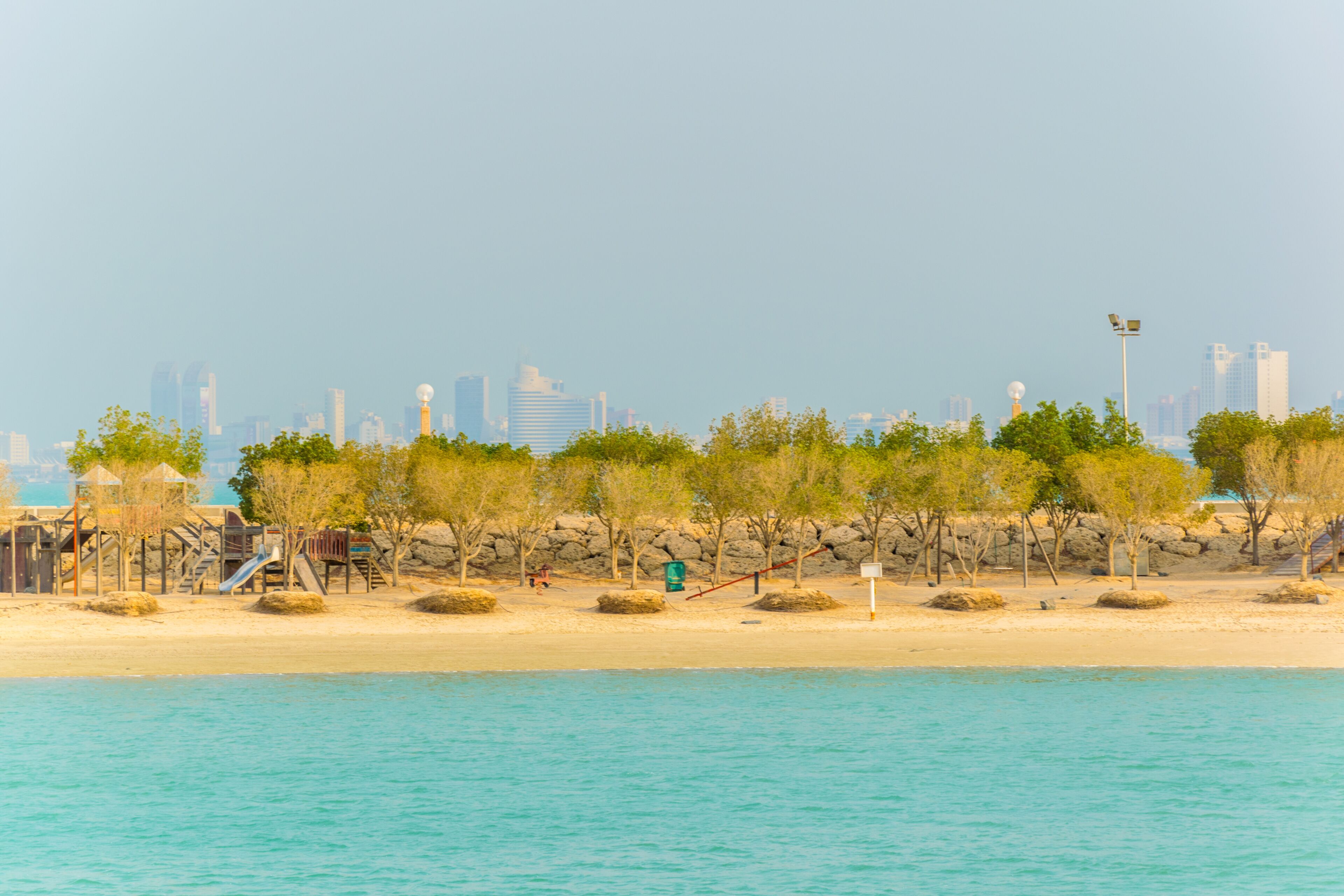 Una playa con un parque infantil, árboles y un horizonte urbano al fondo.