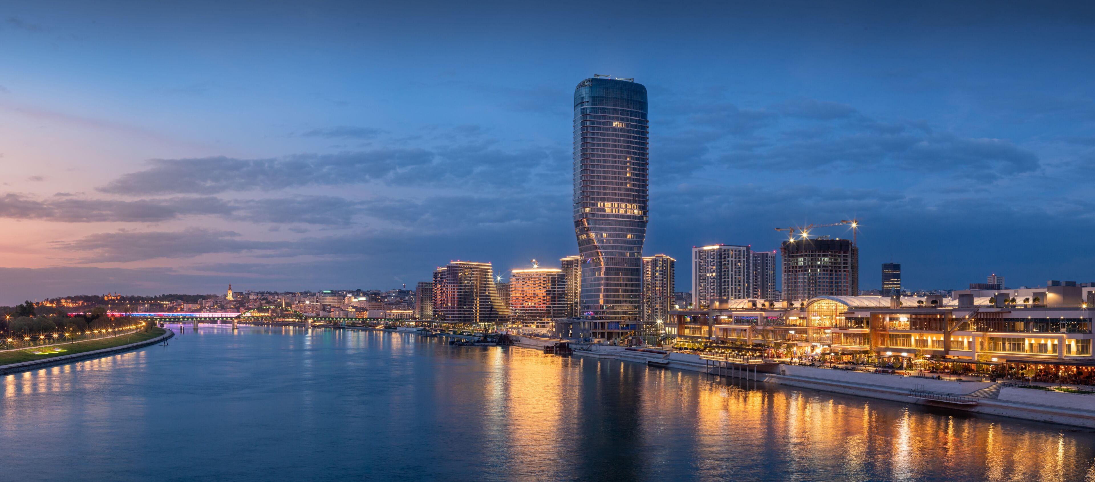 A city skyline with a prominent curved skyscraper, a river reflecting lights, and a bridge in the background.