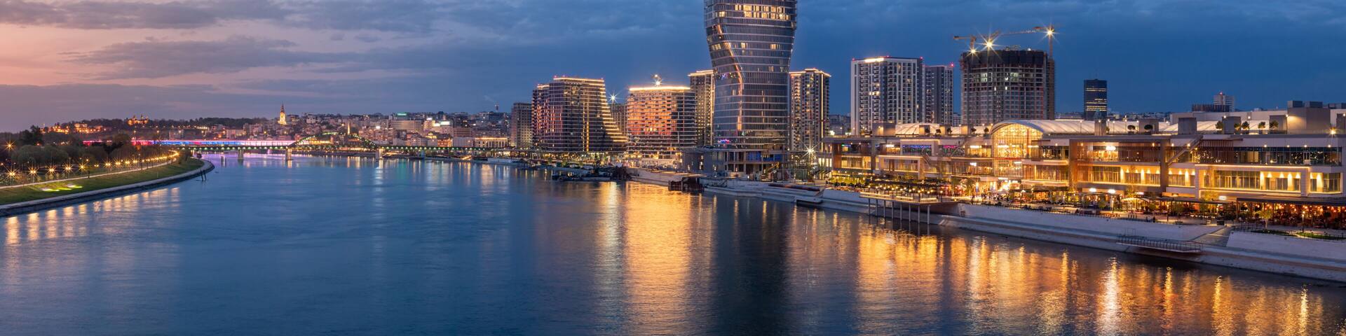 A city skyline with a prominent curved skyscraper, a river reflecting lights, and a bridge in the background.
