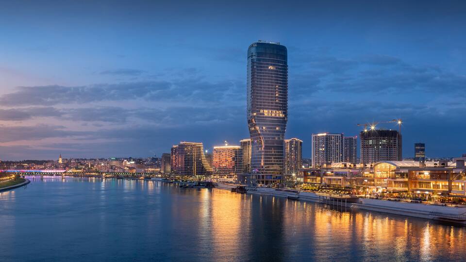 A city skyline with a prominent curved skyscraper, a river reflecting lights, and a bridge in the background.
