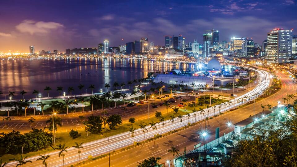 A nighttime cityscape with a waterfront, illuminated buildings, and a curved road with light trails.