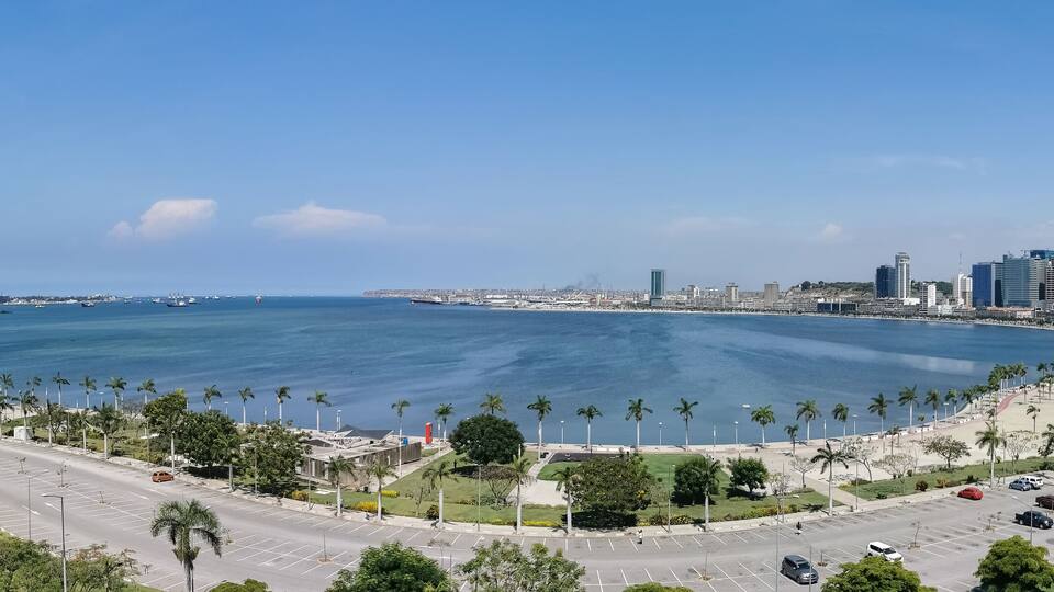 A coastal cityscape with a large body of water, palm-lined streets, and modern high-rise buildings.