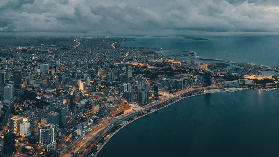 An aerial view of a coastal city with dense urban development, a prominent waterfront, and a curved road leading into the city.