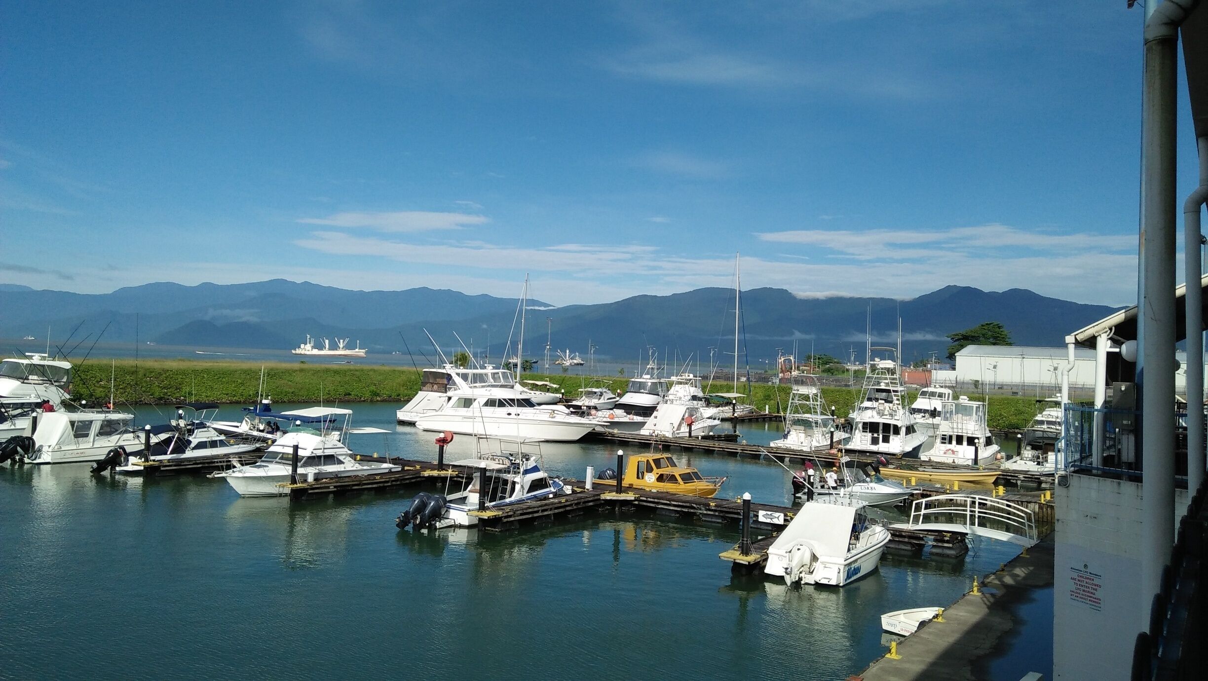 A marina with various boats docked, a large ship in the background, and mountains in the distance.