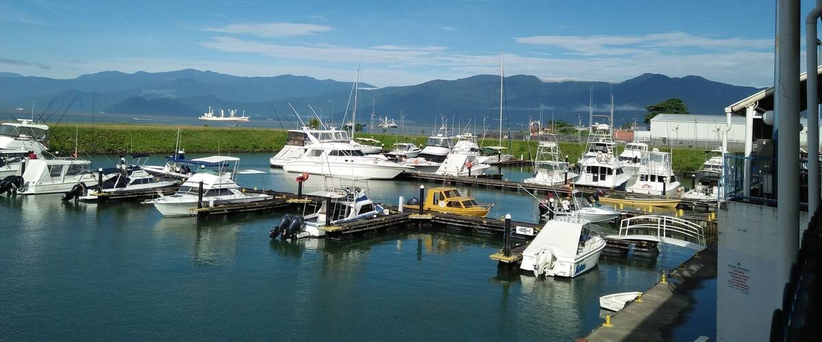 A marina with various boats docked, a large ship in the background, and mountains in the distance.