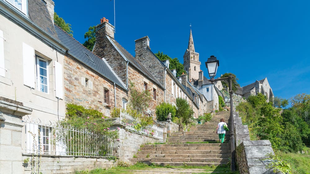 Eine steinerne Treppe führt zu einer Kirche mit einem hohen, schlanken Kirchturm, umgeben von traditionellen Häusern und viel Grün.