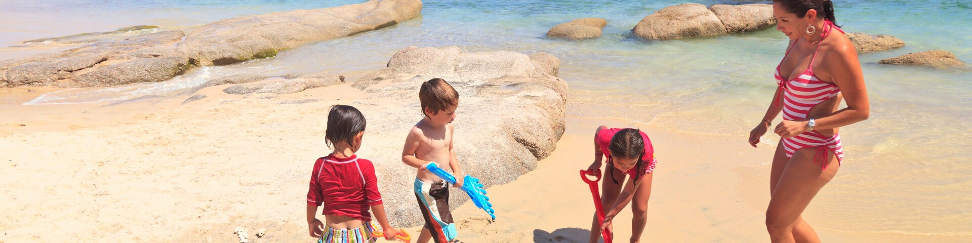 Una playa arenosa con agua azul cristalina, formaciones rocosas y montañas a lo lejos.
