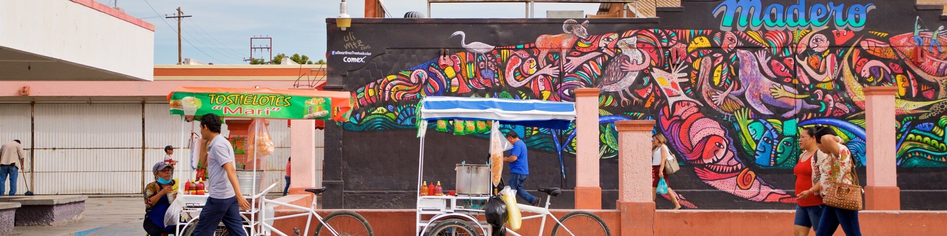 Escena callejera con un mural colorido, un carrito de vendedor ambulante y un letrero que dice 'La Paz B.C. Mercado'.