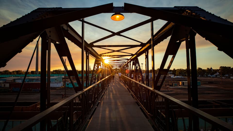A metal bridge with a walkway, metal beams, and a sunset in the background.