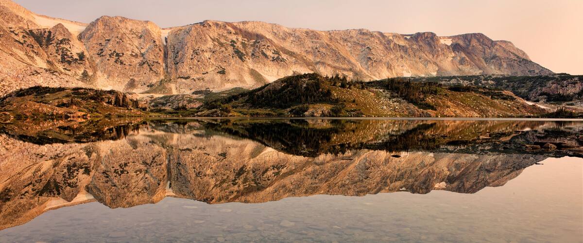 Um lago de montanha com reflexos nítidos, margens rochosas e uma cordilheira arborizada.