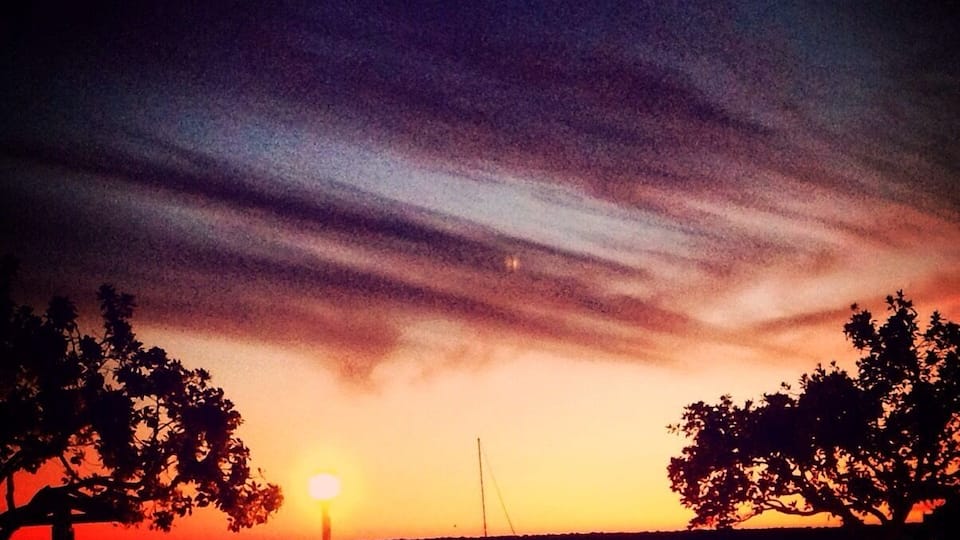 A sunset over a calm body of water with a sailboat, trees, and a dock.