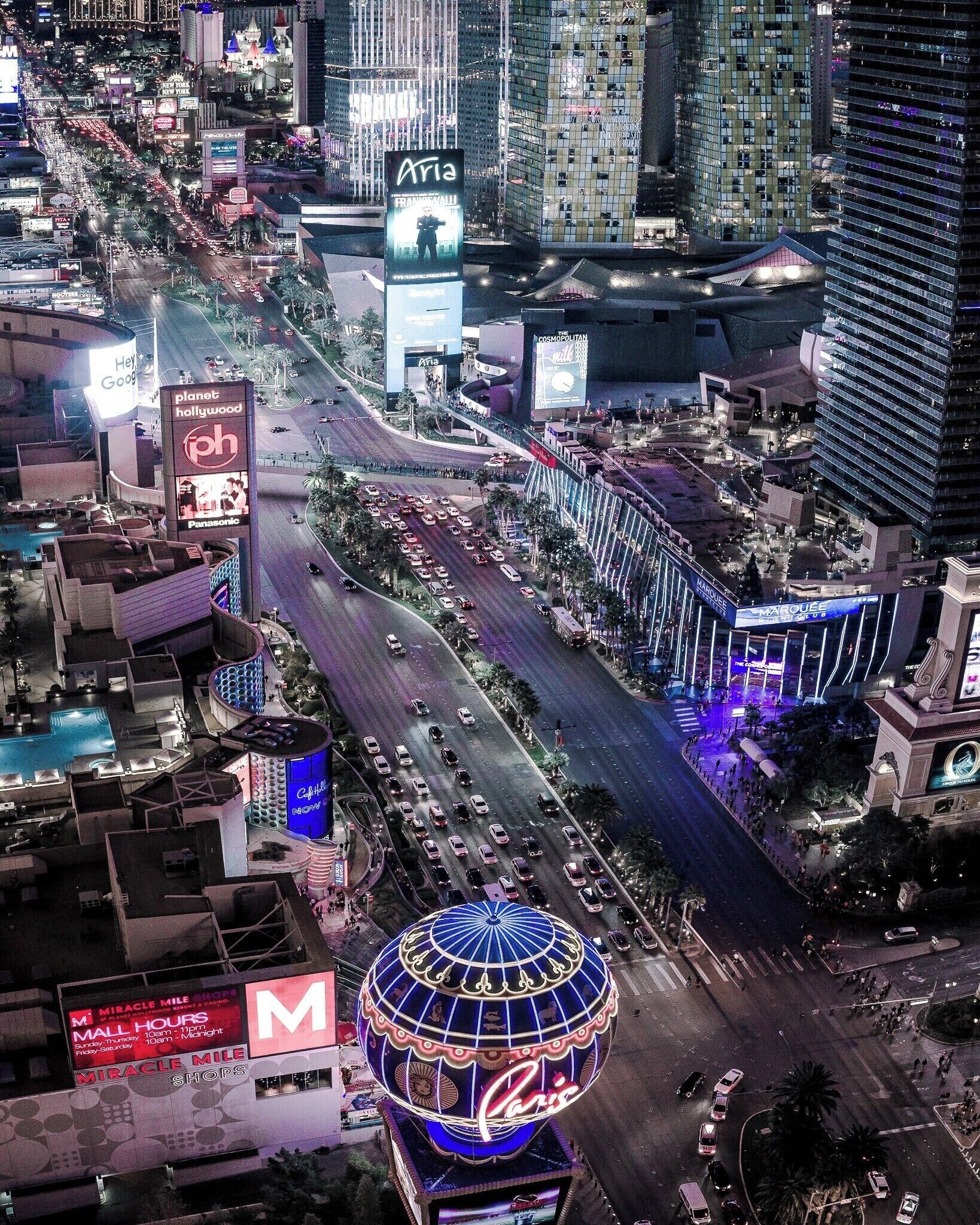 Un paisaje urbano nocturno con pantallas publicitarias iluminadas, una cúpula destacada y un cruce de calles con mucho tráfico.