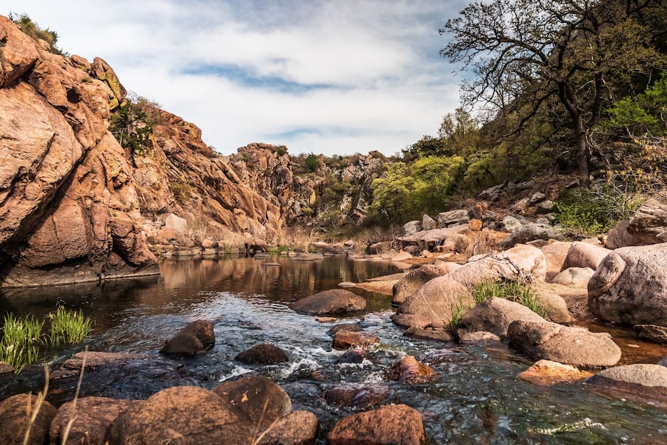 A rocky riverbed with clear water, surrounded by large boulders and sparse vegetation.