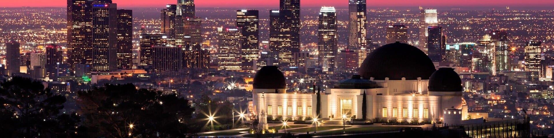 A city skyline at dusk with illuminated buildings and a prominent dome structure in the foreground.