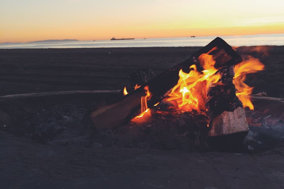Una fogata en la playa al atardecer, con un barco a lo lejos.