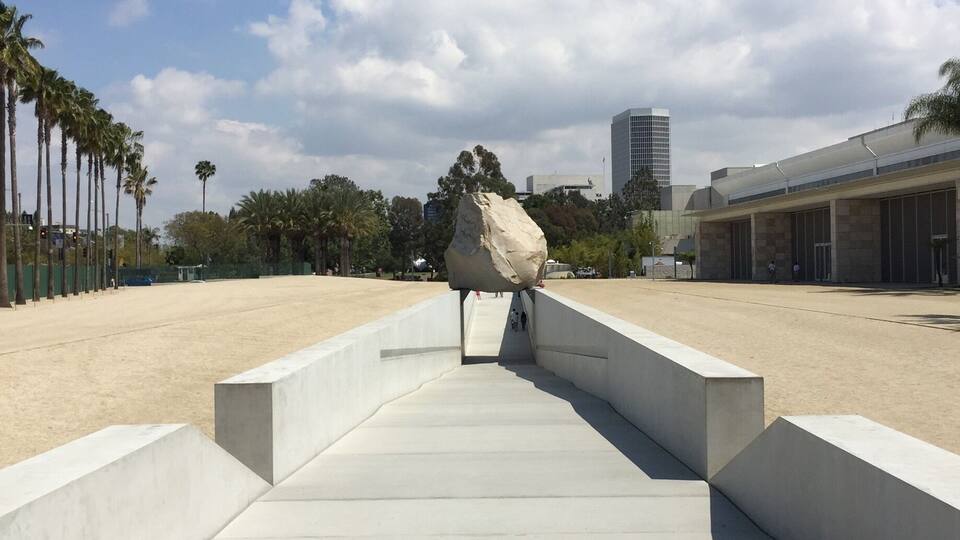 Ein Betonweg führt zu einer großen Felsplastik, Palmen und einem modernen Gebäude im Hintergrund.