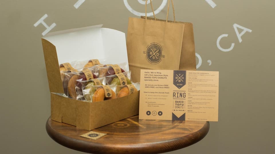 A box of baked tofu donuts, a brown paper bag, and a menu card on a wooden table.