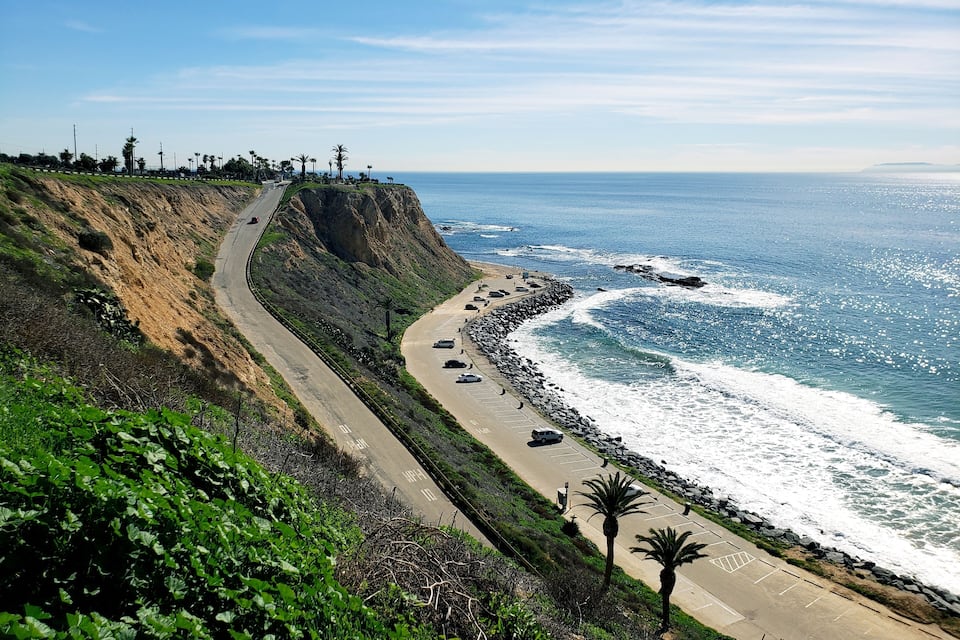 A coastal road with a parking area and a cliff overlooking the ocean.