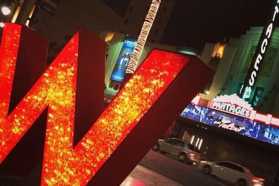Un letrero brillante en forma de 'W' con un resplandor neón, un letrero de 'PANTAGES' y un edificio con señalización iluminada al fondo.