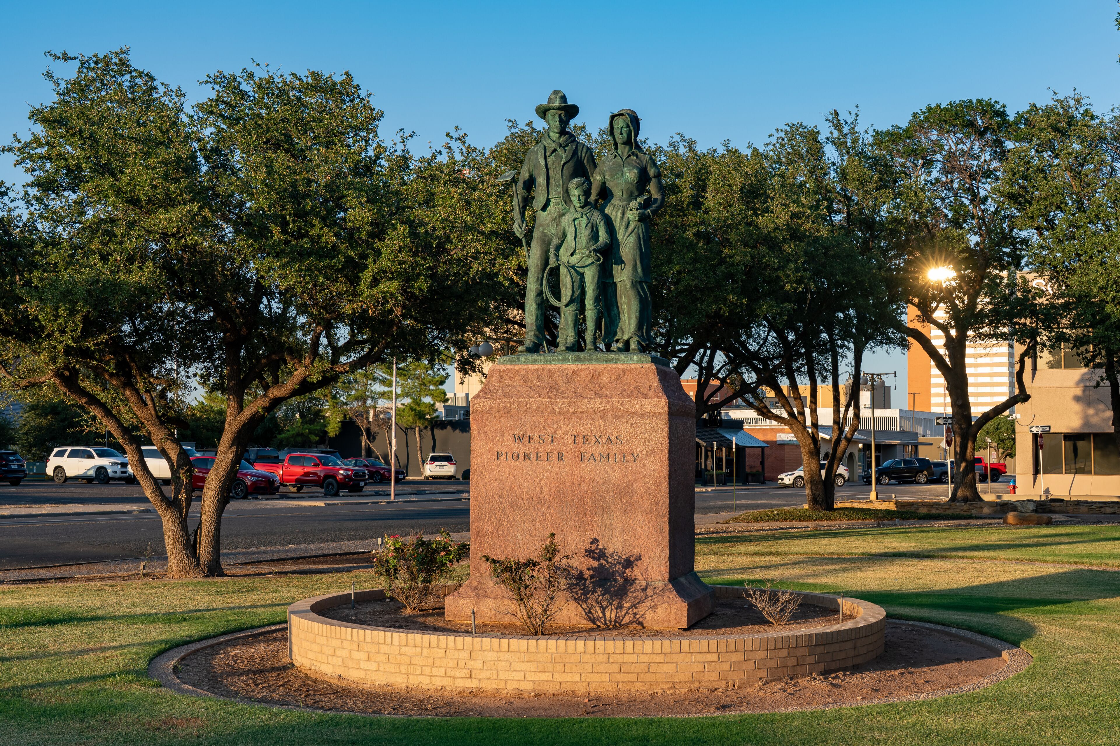 A bronze statue of a family with a plaque reading 'West Texas Pioneer Family' in a landscaped park with trees and a road in the background.