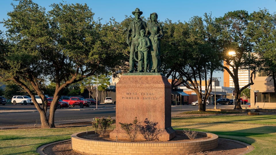 A bronze statue of a family with a plaque reading 'West Texas Pioneer Family' in a landscaped park with trees and a road in the background.