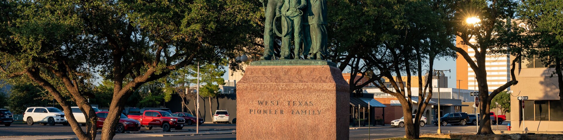 Una estatua de bronce muestra a una familia con una placa que dice 'West Texas Pioneer Family' en un parque arbolado, con un camino al fondo.