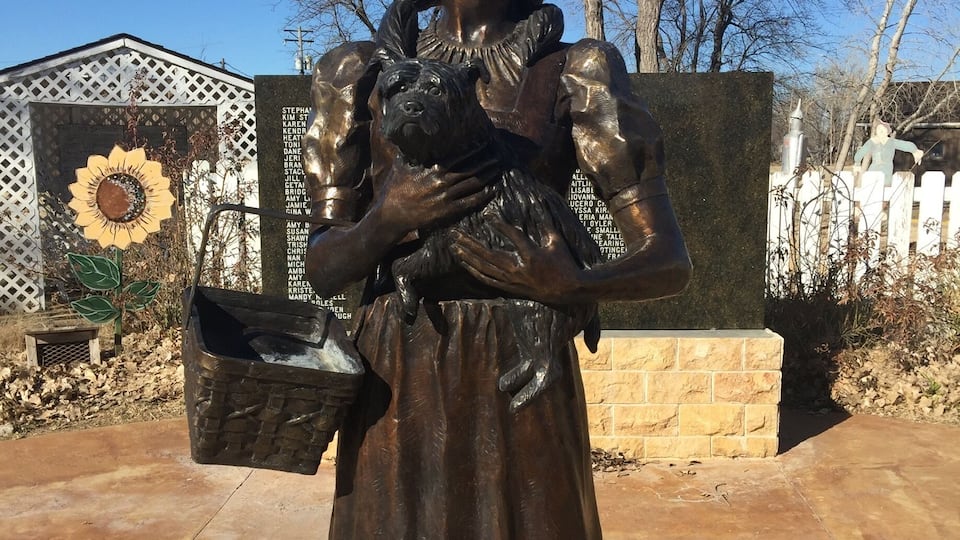 A bronze statue of a woman holding a dog, with a sunflower and a fence in the background.