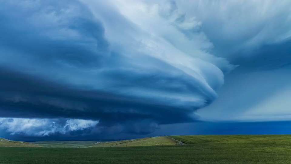A vast landscape with rolling green hills and a dramatic sky featuring a large, swirling cloud formation.