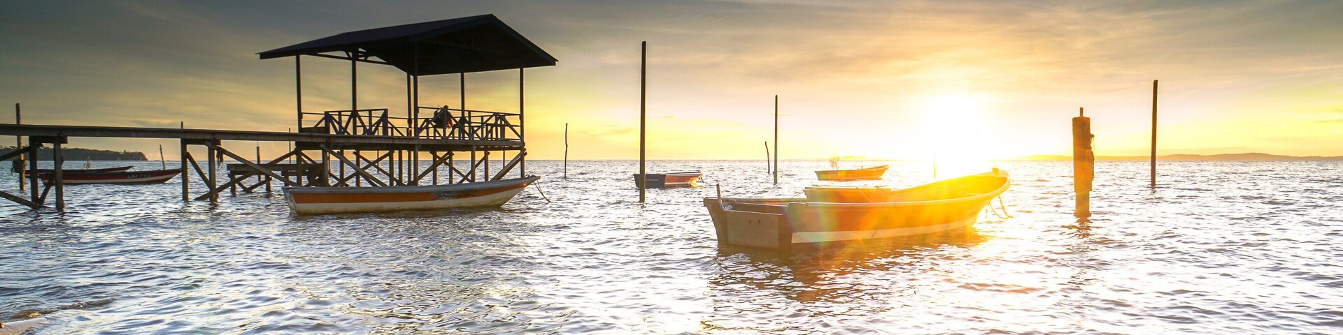 Un muelle con una zona de estar cubierta y un barco fondeado al atardecer.
