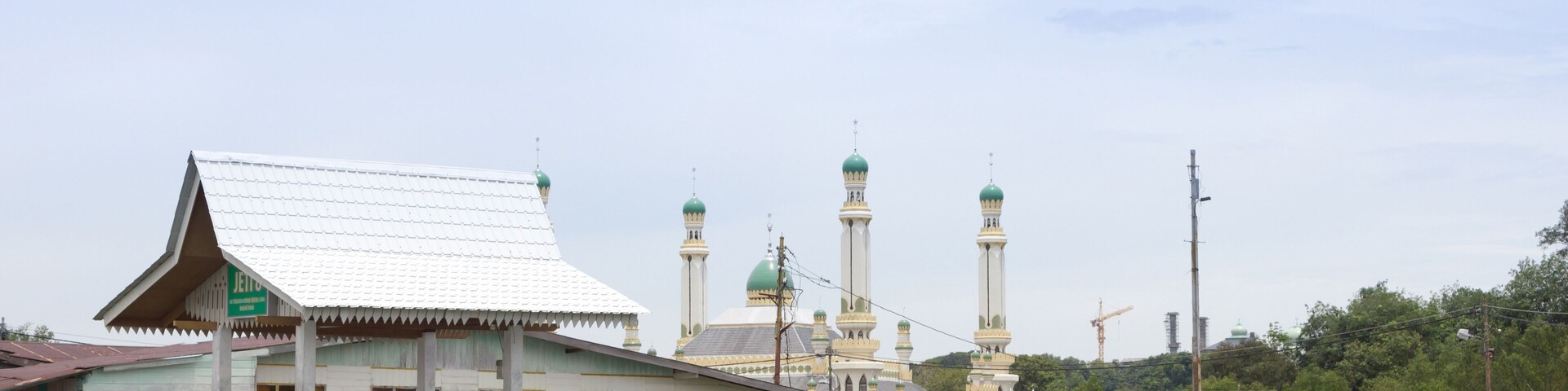 Uma mesquita com cúpulas verdes e minaretes, passarelas de madeira sobre a água, e uma grua de construção ao fundo.
