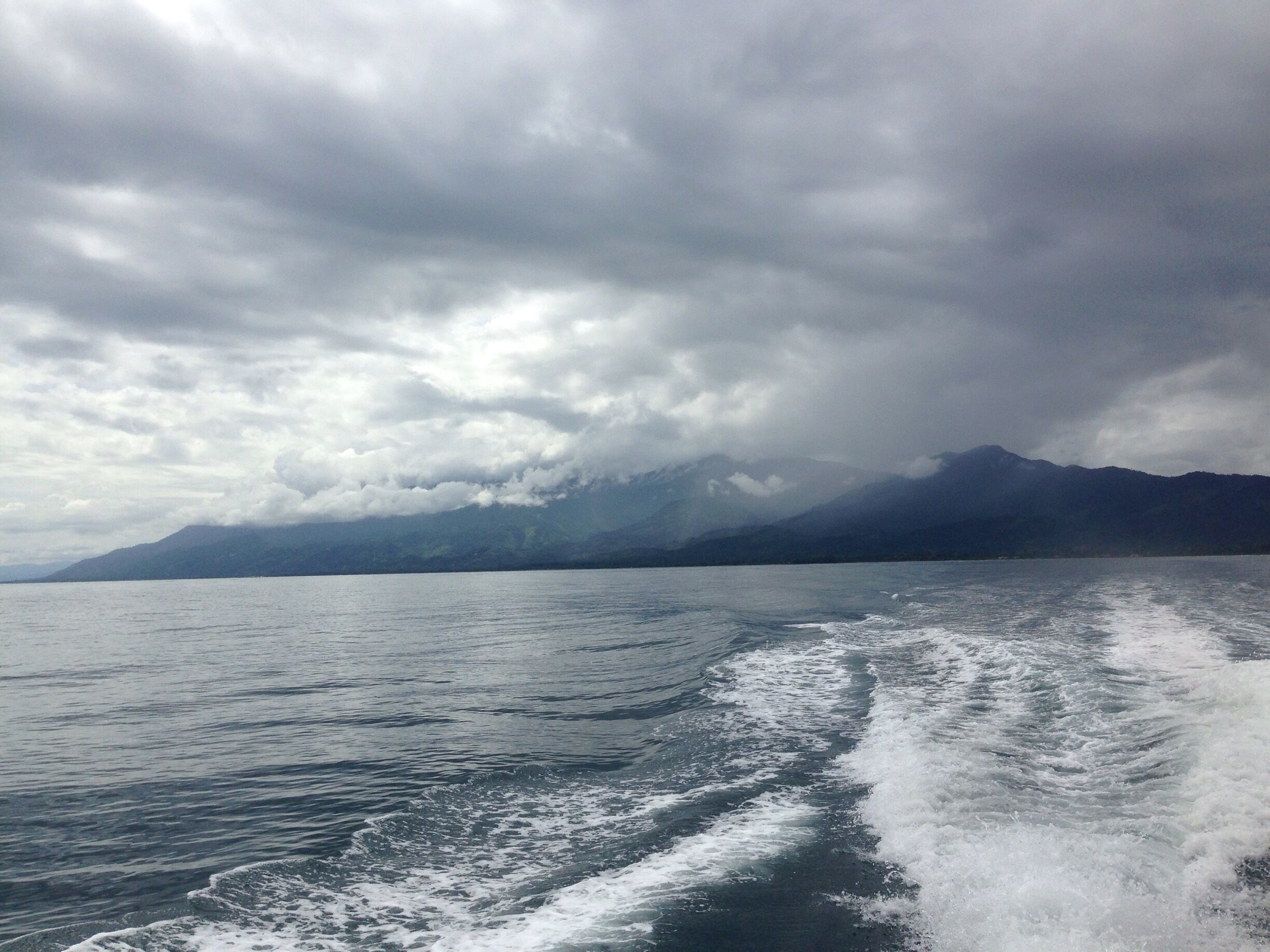 A body of water with waves, a mountain range in the distance, and a cloudy sky.