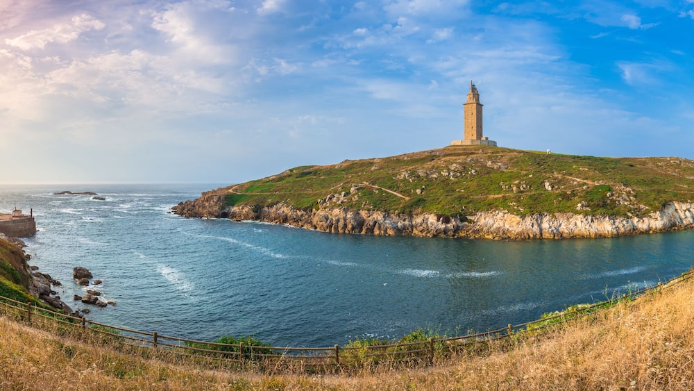 Un paysage côtier avec un phare perché sur une colline, une plage et une côte rocheuse.