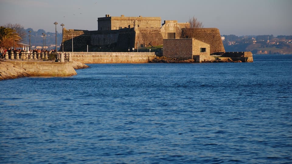 A historic stone fortress by a calm sea with a promenade and distant hills.