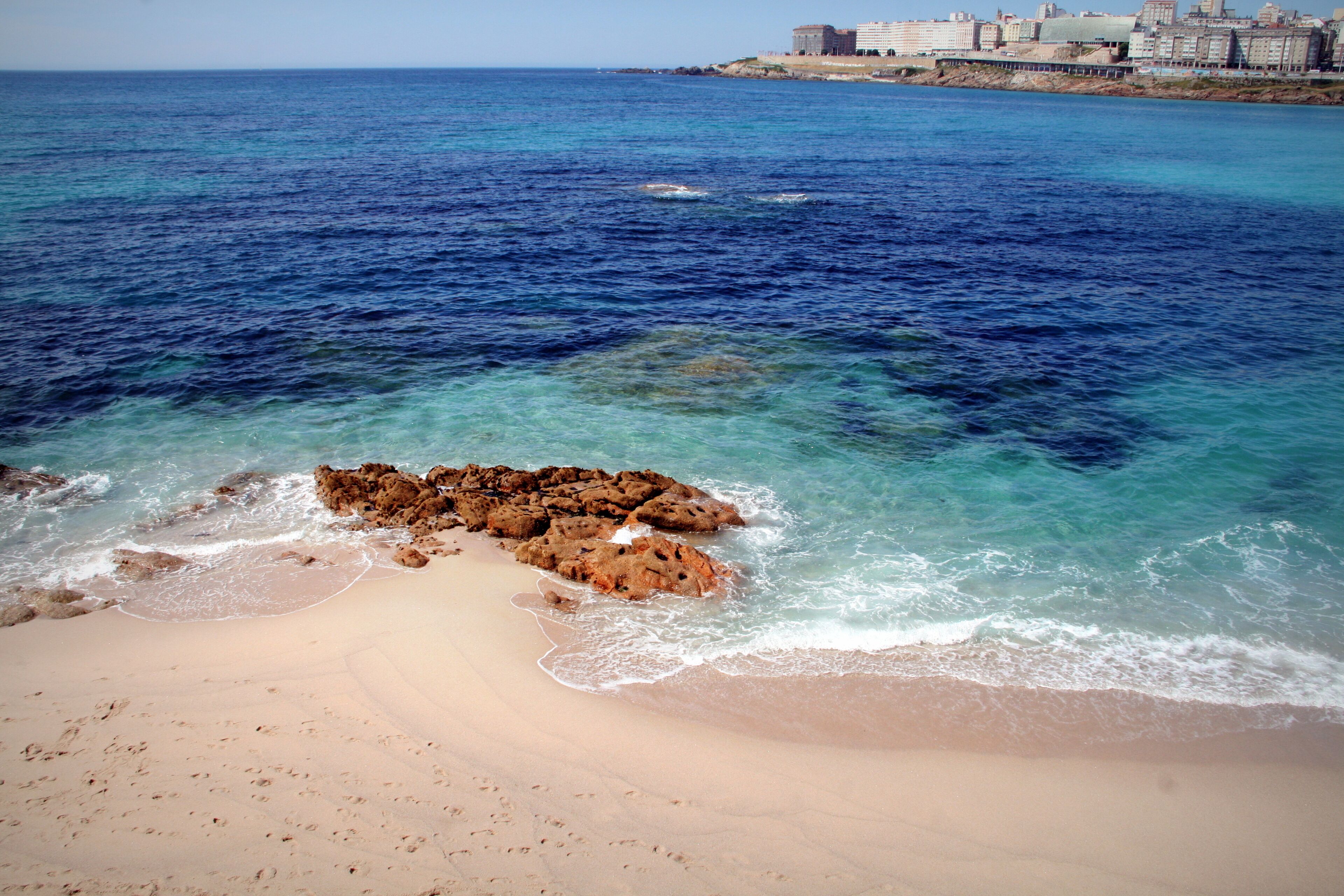 A beach with clear turquoise water, a rocky outcrop, and a cityscape in the background.