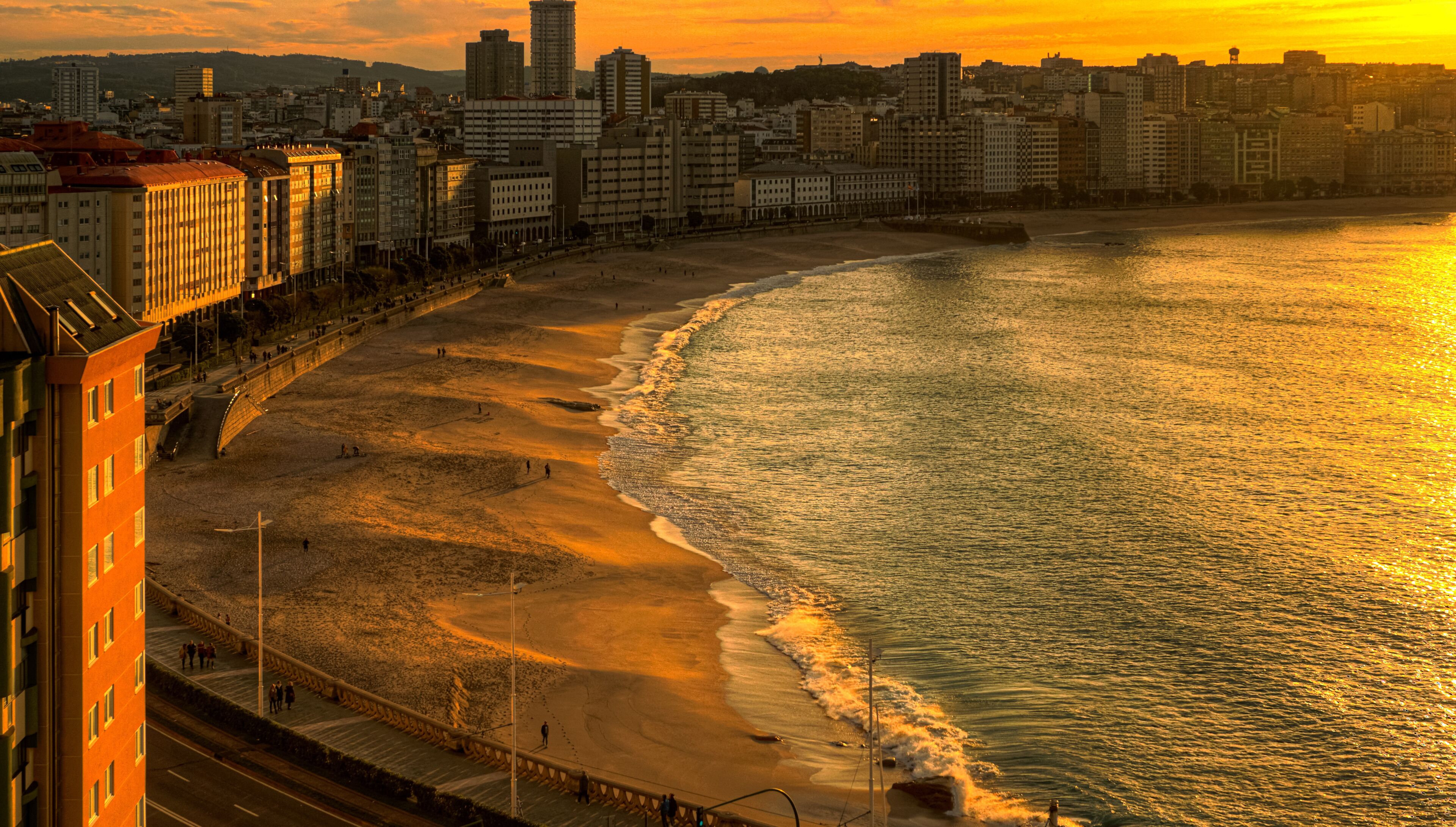 A coastal cityscape with a wide beach, a promenade, and high-rise buildings under a sunset sky.