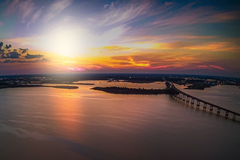 A sunset over a calm body of water with a bridge and distant city lights.