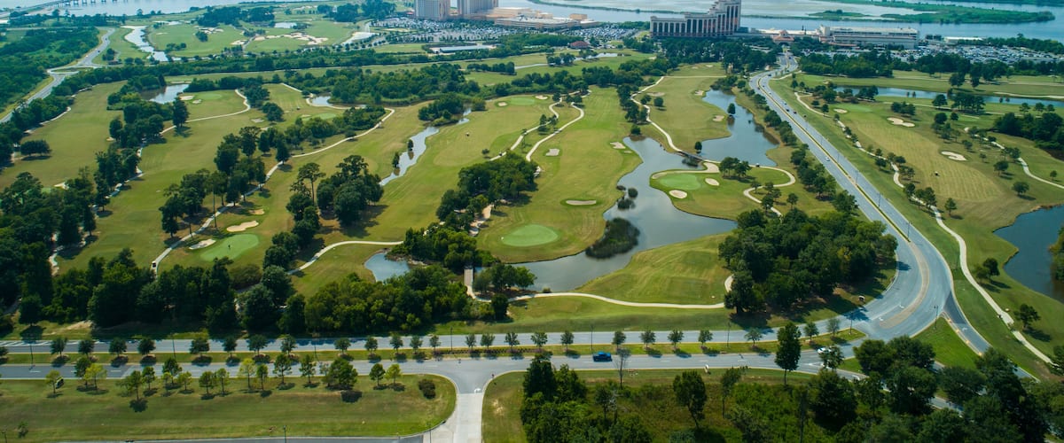 An aerial view of a golf course with multiple water hazards, a bridge, and surrounding buildings.